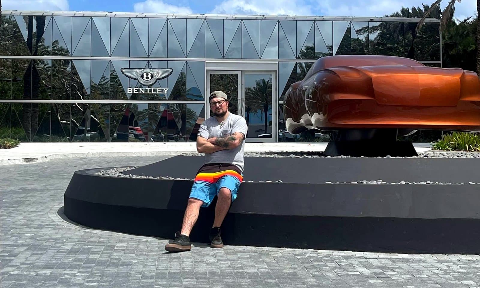 Sam assis devant un studio de design Bentley avec une sculpture de voiture